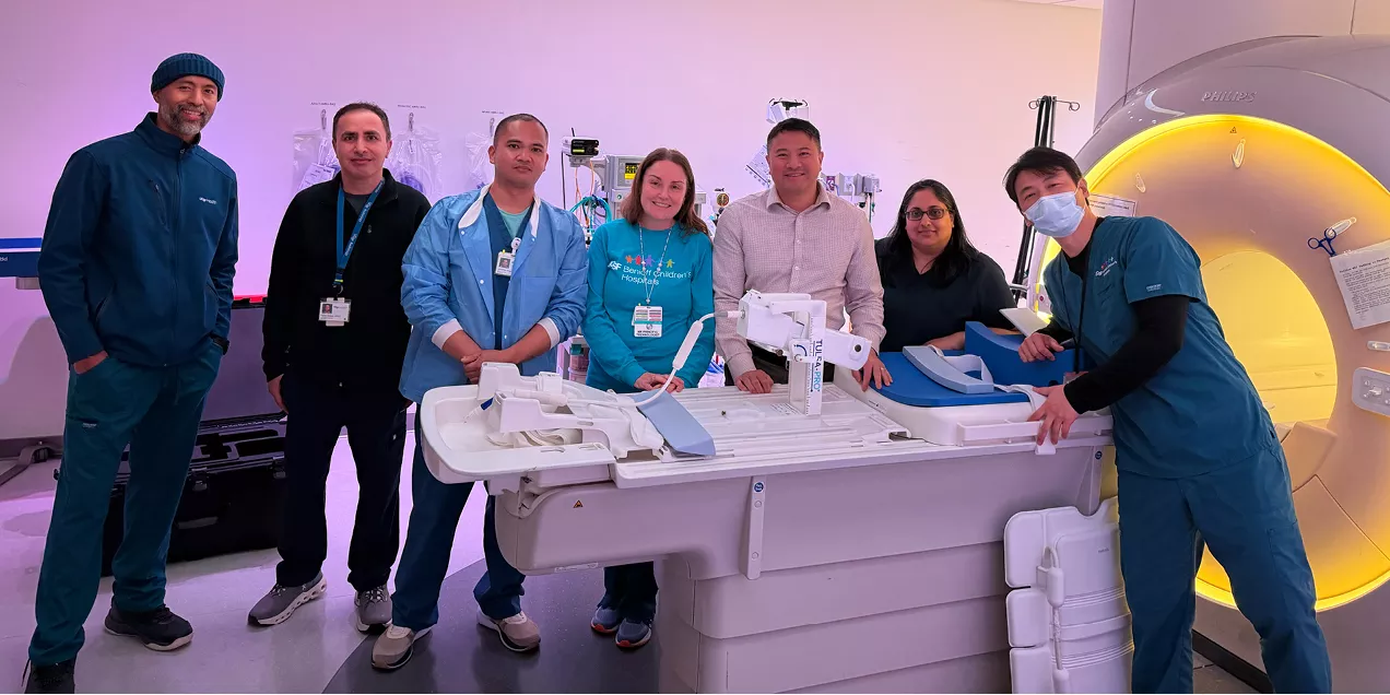 Group of seven healthcare professionals standing around an MRI scanner in a clinical imaging suite, posing together beside the patient table and scanner.