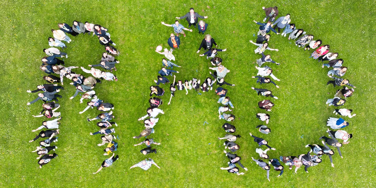 Aerial view of dozens of people lying on a grassy lawn to spell "RAD," with a few people standing in the center and flowering trees visible at the edge of the image.