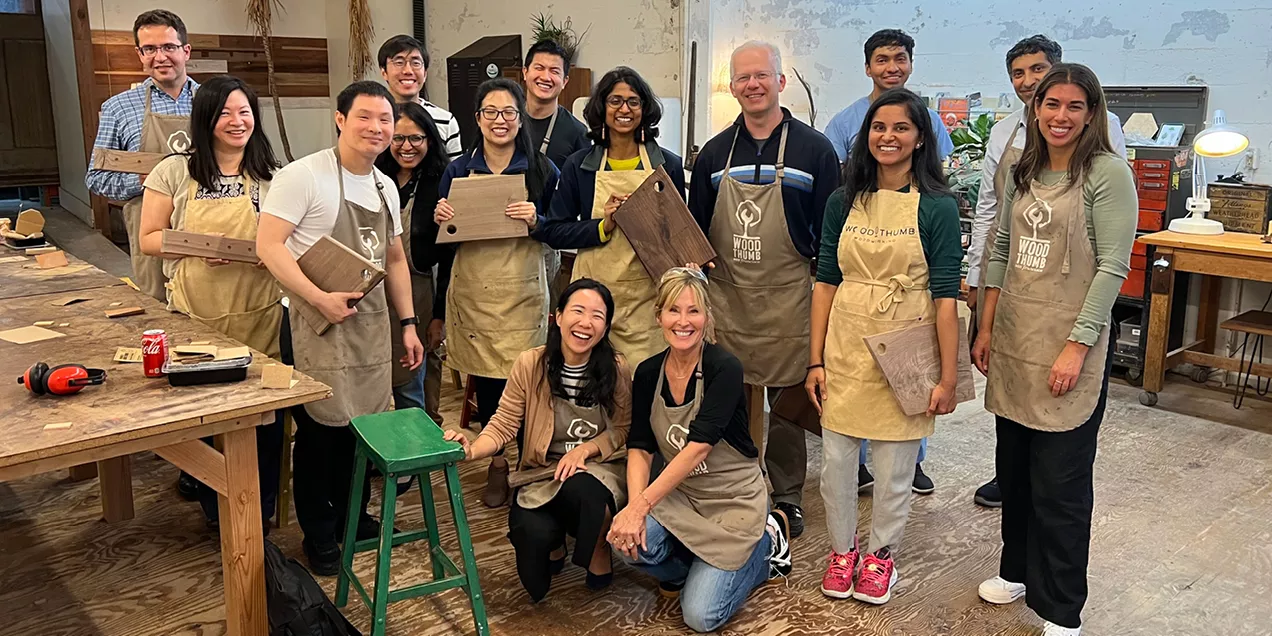 A group of 12 people wearing tan woodworking aprons pose together inside a woodworking studio, smiling and holding handmade wooden cutting boards. Two people kneel in the front while the others stand behind them. Workbenches, tools, unfinished wood projects, and workshop equipment are visible in the background, highlighting a collaborative team-building woodworking event.