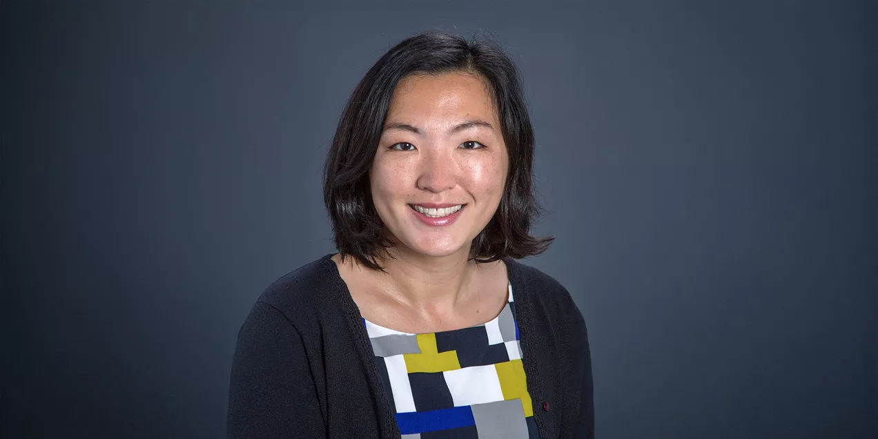 A professional head-and-shoulders portrait of a smiling person against a solid dark gray background. She has shoulder-length dark hair and is wearing a black cardigan over a geometric-patterned blouse in white, gray, black, yellow, and blue. She faces the camera with a friendly expression in a studio-style photograph.