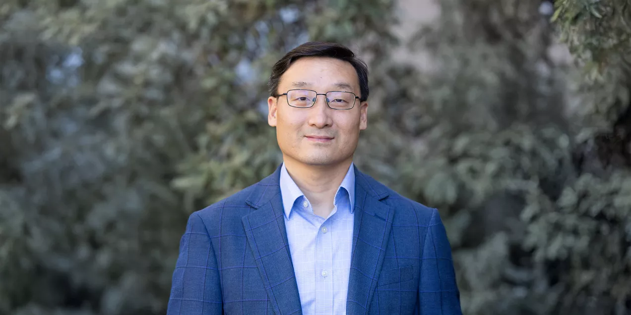 Portrait of a man wearing glasses, a blue blazer, and a light blue dress shirt, standing outdoors in front of softly blurred evergreen trees and looking at the camera with a slight smile.