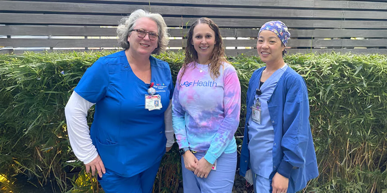 Three nurses stand outdoors in front of greenery, smiling at the camera. The nurse on the left wears blue scrubs with an ID badge, the person in the center wears a colorful UCSF Health tie-dye sweatshirt and light-colored pants, and the nurse on the right wears blue scrubs with a patterned surgical cap. The group poses together in recognition of Certified Nurses Day.