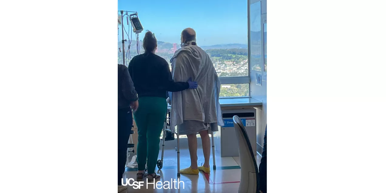 Patient wearing a neck brace stands with a walker beside a caregiver, looking out a hospital window at the Golden Gate Bridge during rehabilitation.