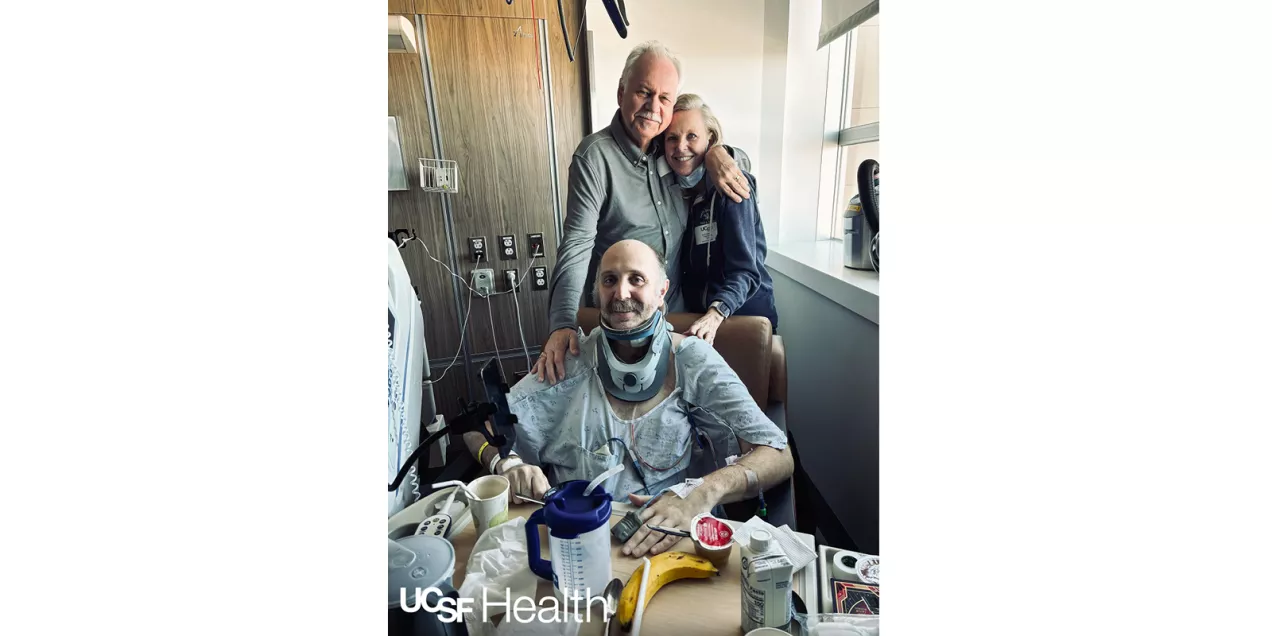 A smiling patient wearing a neck brace sits in a recliner at home with two young children wearing bunny ears, sharing a joyful family moment.