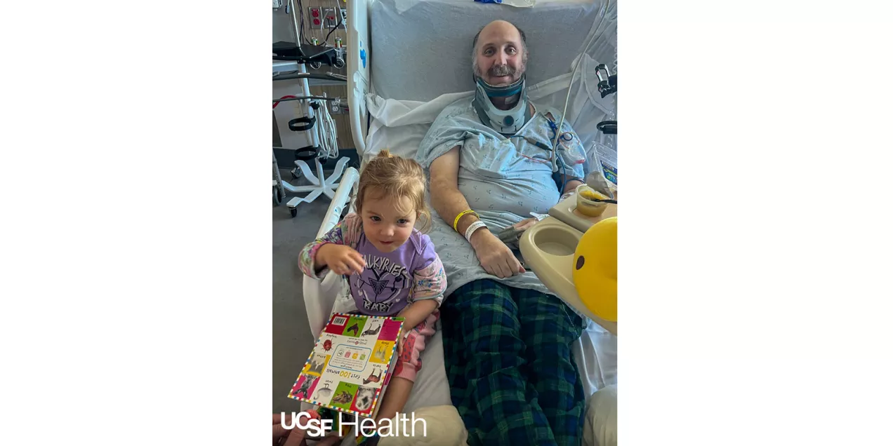 A patient wearing a neck brace reads a children's book with a young child curled up beside him, surrounded by a stack of books at home.