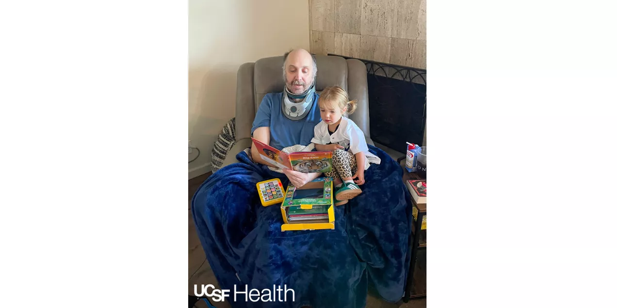 A hospitalized patient wearing a neck brace smiles from a hospital bed while a young child sits beside him holding a picture book.