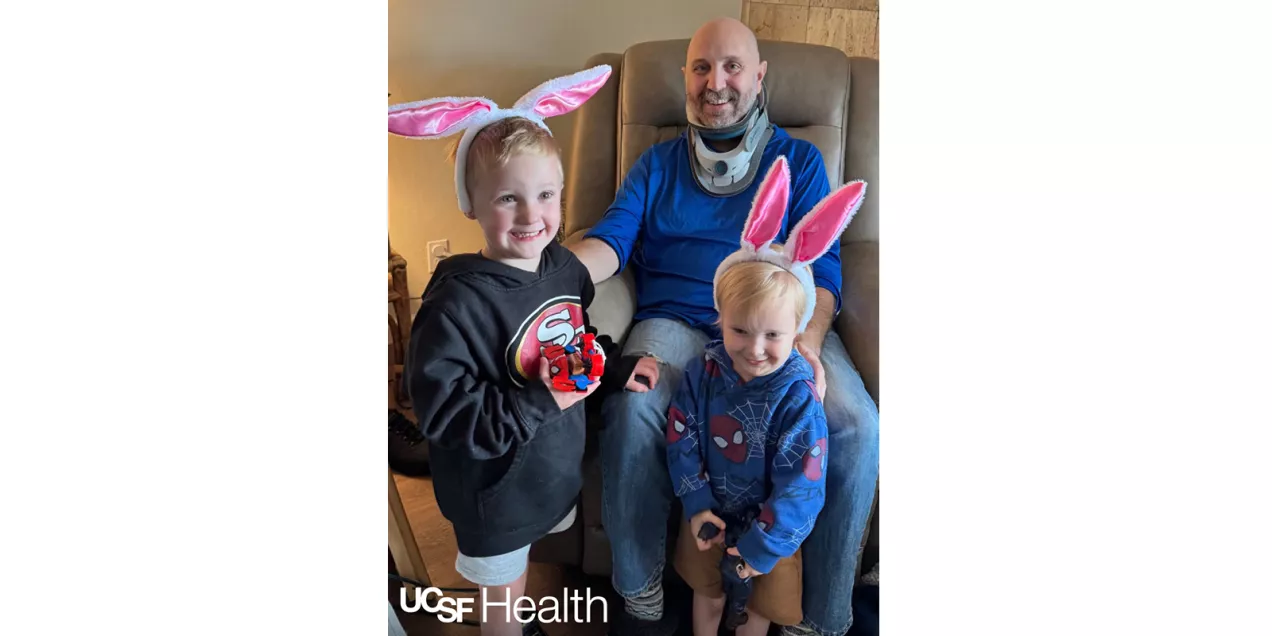 A patient wearing a neck brace smiles while seated in a hospital room with two supportive family members standing behind him, embracing him for a family portrait.