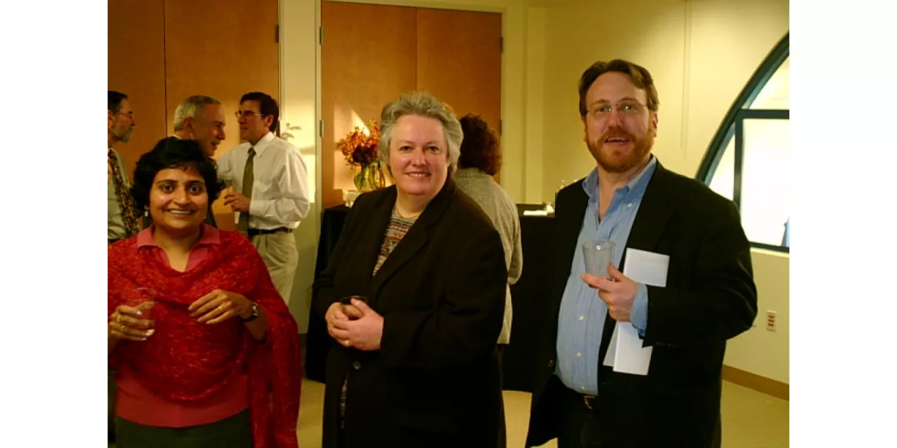 Three colleagues gather at an indoor reception, with other attendees conversing in the background