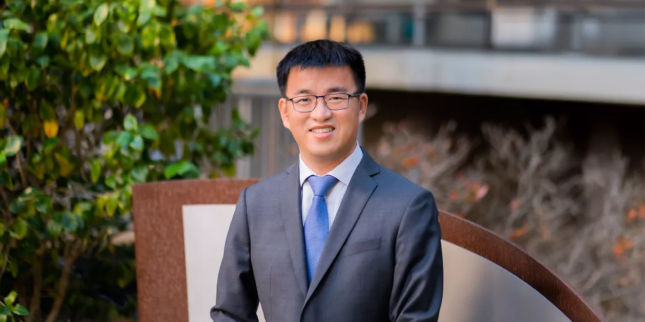 Professional portrait of a smiling man wearing glasses, a gray suit, white shirt, and blue tie, standing outdoors in front of greenery and a blurred building.