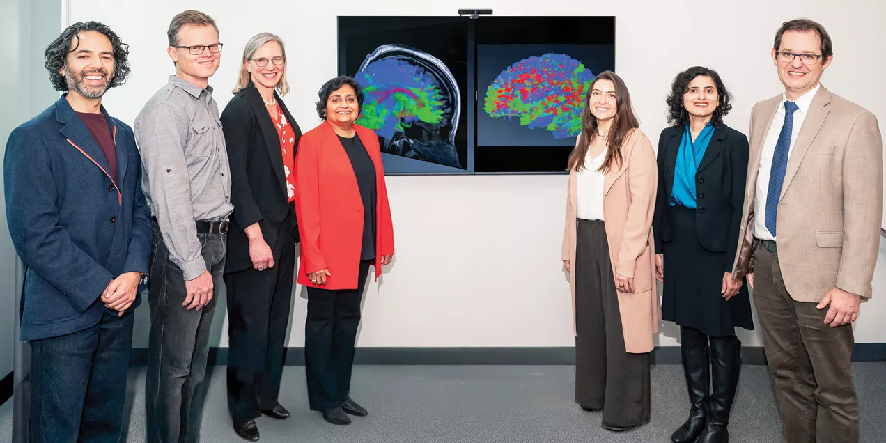 Seven people stand together in a clinical or research setting beside two wall-mounted monitors displaying colorful brain imaging scans.