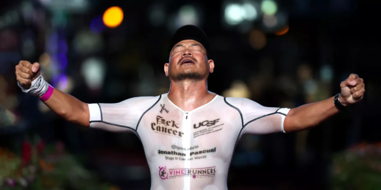 Athlete crossing a finish line at night with arms raised in celebration, wearing a white racing suit displaying cancer awareness and UCSF-related logos.