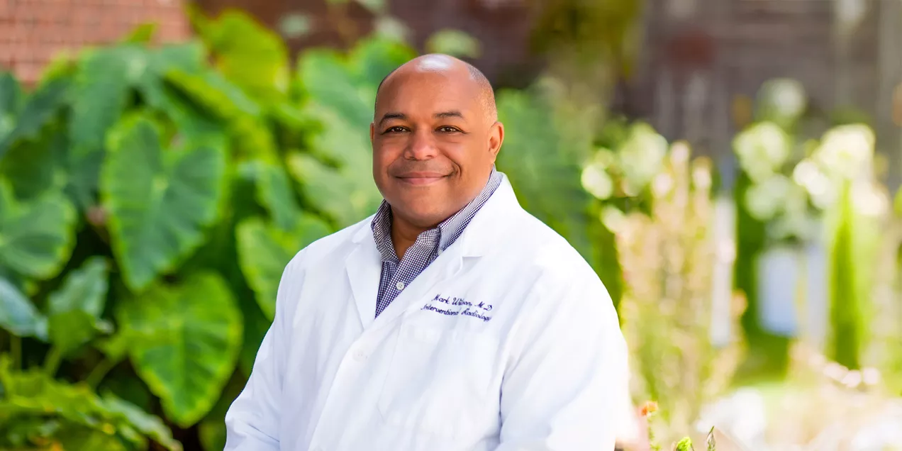 Portrait of a physician wearing a white coat outdoors against a backdrop of green foliage.