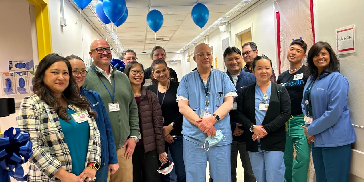A group of healthcare staff pose together in a hospital hallway during a ribbon-cutting celebration. Blue balloons and smiles mark the occasion, with clinicians and staff wearing a mix of scrubs, lab coats, and business attire.