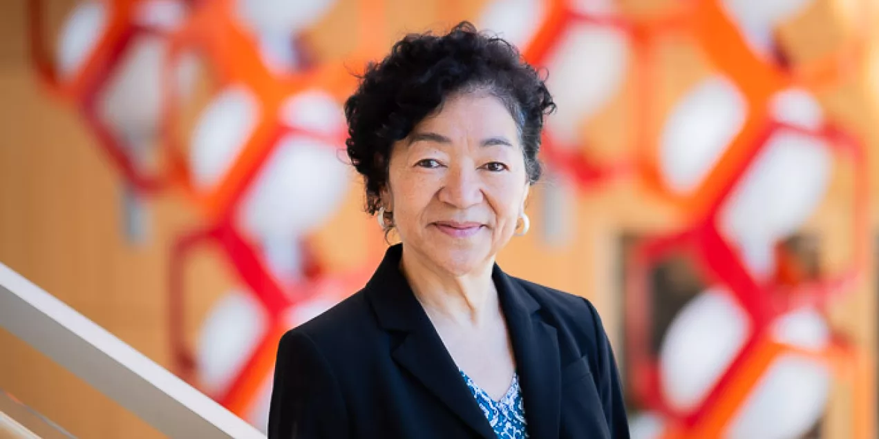 A woman in a dark blazer smiles at the camera, standing in front of a colorful red and orange geometric background.