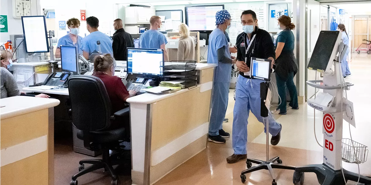 Healthcare workers in scrubs and masks work around a busy hospital nursing station with computer workstations and medical equipment.
