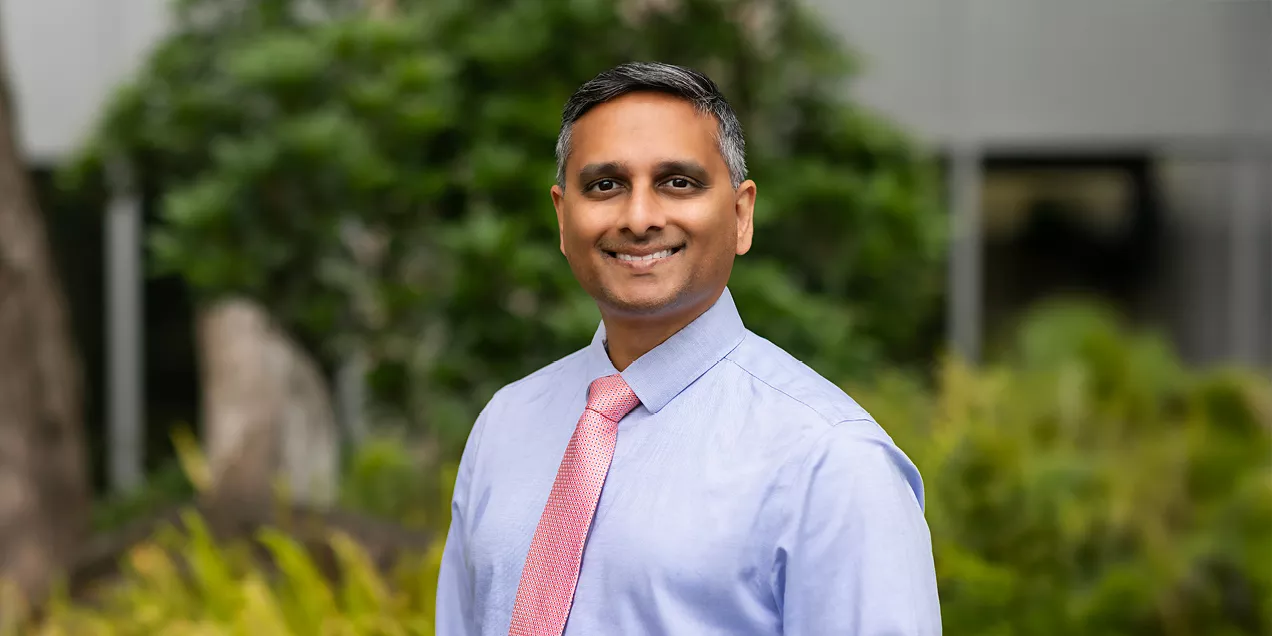Portrait of a smiling man wearing a light blue dress shirt and coral tie, standing outdoors with greenery in the background.
