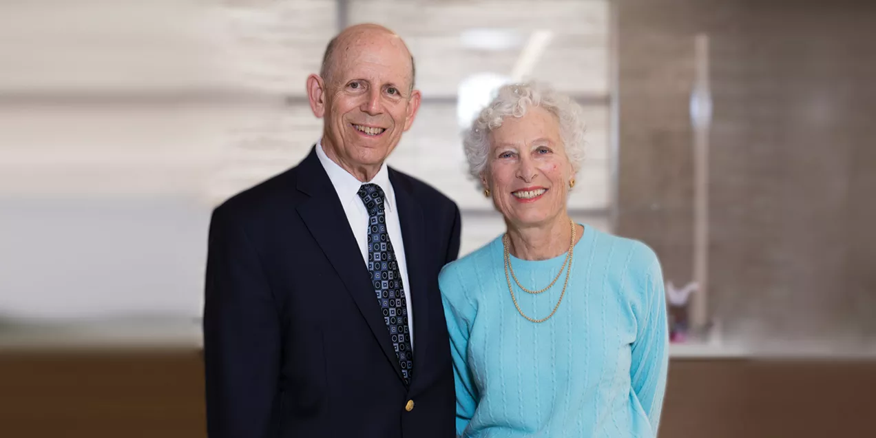 An older man in a dark suit and tie stands beside an older woman wearing a light blue top and gold necklaces. Both are smiling at the camera.