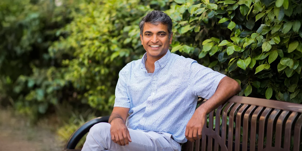 Man smiling while seated on a bench outdoors surrounded by greenery.