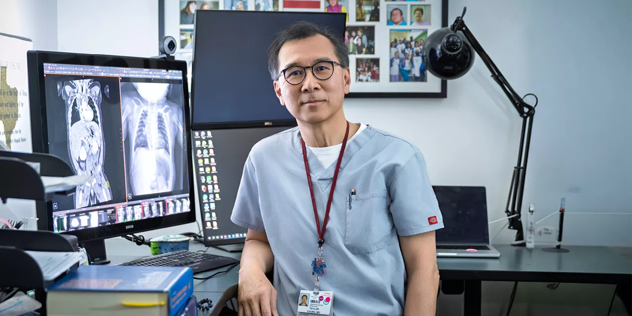 Man wearing medical scrubs and glasses seated at a workstation with medical images displayed on a monitor.