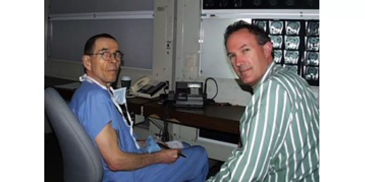 Two men seated in a radiology reading room with medical images displayed in the background.
