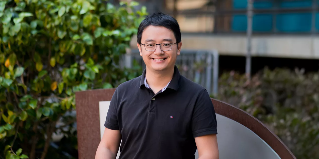 Man wearing glasses and a black polo shirt smiling outdoors with greenery in the background.