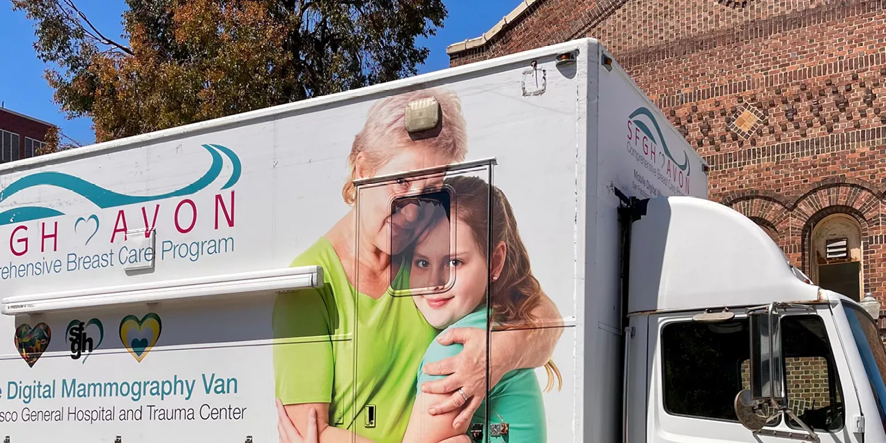 Mobile breast care and mammography van parked beside a brick building, featuring a large photograph of an older woman embracing a young girl on its side.