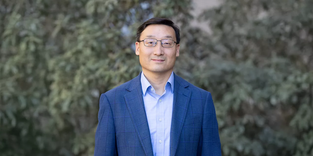 Portrait of a man wearing glasses, a blue blazer, and a light blue shirt, standing outdoors against a softly blurred background of trees and greenery.