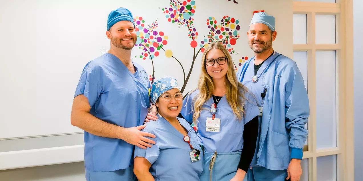 Group photo of four healthcare professionals standing together in a hospital or clinical setting. The team is dressed in blue medical scrubs, with two members also wearing surgical caps. They are smiling toward the camera and standing in front of a colorful wall mural depicting a tree made of multicolored circular leaves. Identification badges are visible on several team members, and one person has a hand resting on a colleague’s shoulder. The image conveys teamwork, compassion, and a supportive healthcare 