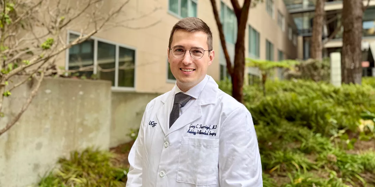 A man wearing glasses, a white UCSF medical coat, dress shirt, and tie stands outdoors on a landscaped medical campus, smiling at the camera.