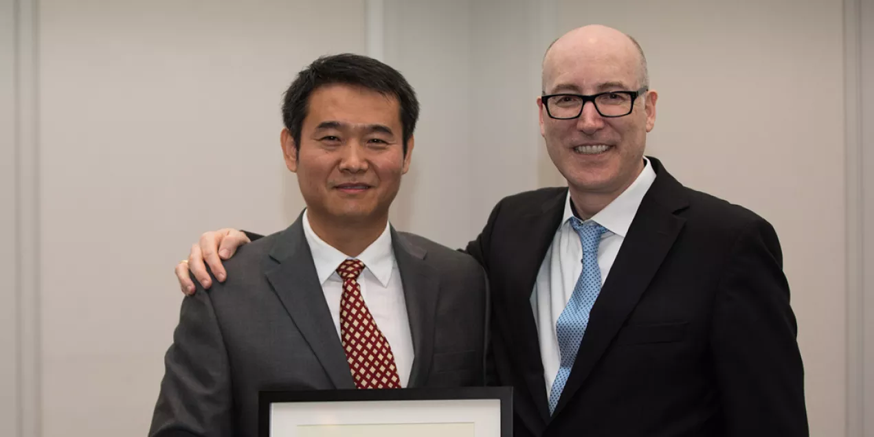Two people in suits standing together at an award event, smiling and holding a framed certificate. One has an arm around the other in a congratulatory pose, and the setting appears to be an indoor formal venue.