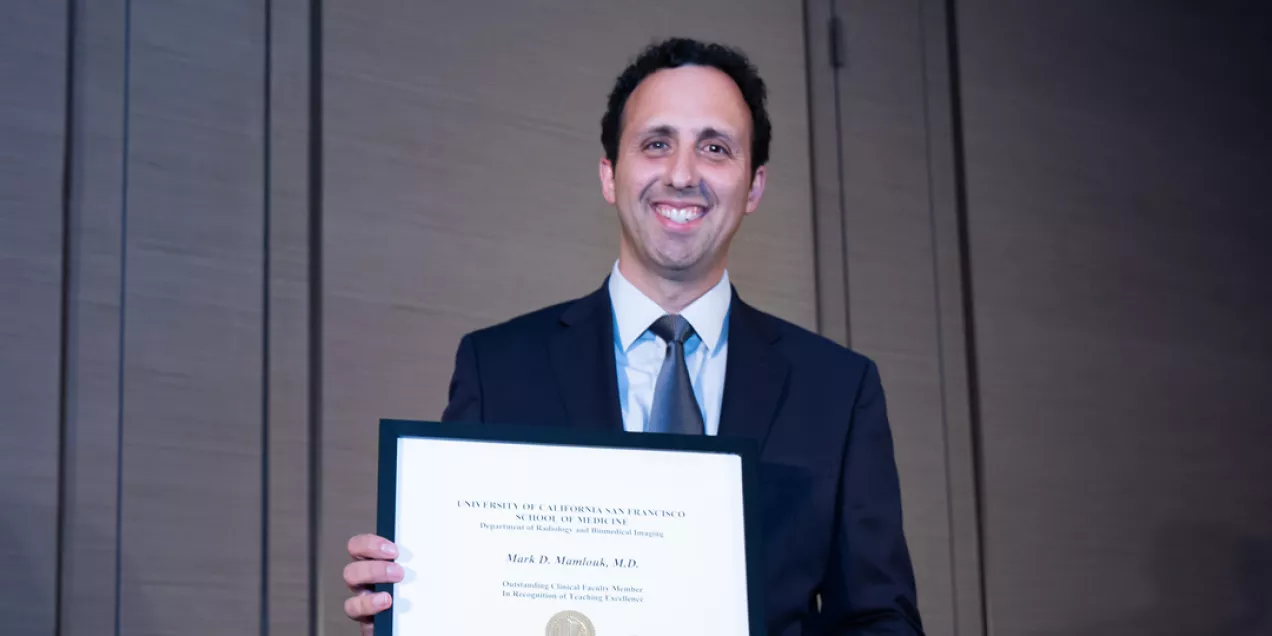 Mark Mamlouk smiling while holding a framed certificate at an indoor award event, standing against a paneled wall.