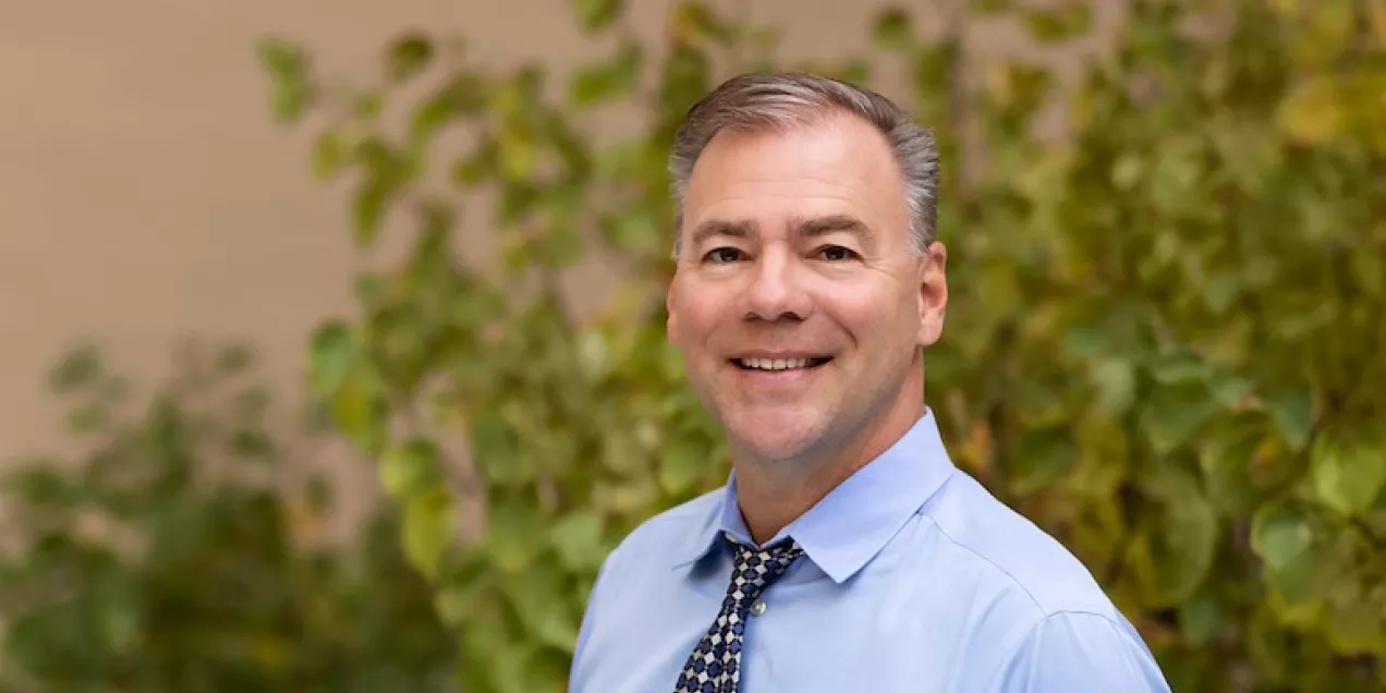 Jeff Geiger smiling outdoors, standing in front of bright green foliage with soft natural light in the background.