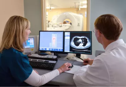 two clinicians in a control room reviewing CT scan images on dual monitors while a patient undergoes a scan in a nearby imaging suite visible through the window