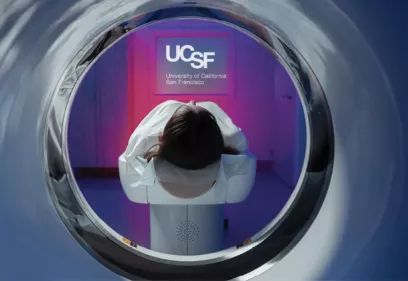 View from inside an MRI scanner looking outward at a patient lying on the table, facing a wall lit with purple and blue light and a UCSF logo projected in front of them.