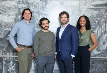 Four people standing side by side and smiling in front of a large, detailed grayscale mural depicting a cityscape and historical scenes.