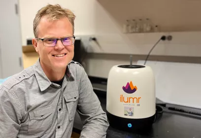 Pedar Larson seated in a lab next to a small tabletop MRI device labeled ilumr, smiling toward the camera in a workspace with equipment in the background