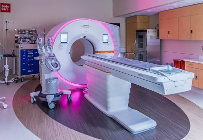 Photon-counting CT imaging suite with a Siemens scanner glowing with pink accent lighting, surrounded by medical equipment, cabinets, and a patient table in a clean clinical room