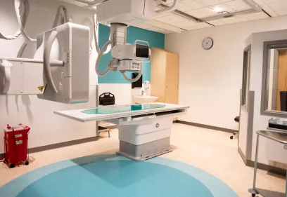 x-ray imaging room with a large radiography table, ceiling-mounted x-ray tube, detector panel, and surrounding medical equipment, shown in a clean clinical setting with teal and white walls