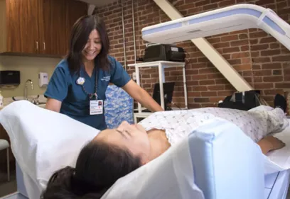 technologist assisting a patient lying on a padded table during a body composition x-ray scan, with the scanning arm positioned overhead in a clinical room with brick walls