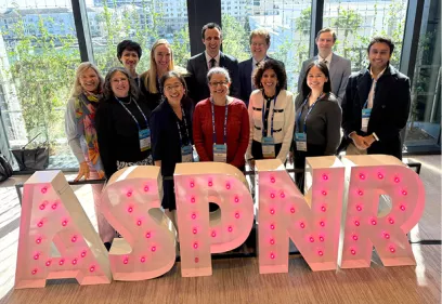 Group photo of UCSF faculty, alumni, and trainees standing indoors behind large illuminated ASPNR letters, smiling at the camera, with conference badges visible and a city view through floor to ceiling windows in the background.