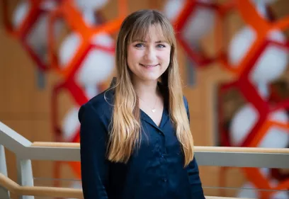 A woman with long blonde hair and bangs wearing a dark blouse stands indoors, smiling at the camera, with a softly blurred background featuring red geometric structures and warm tones.