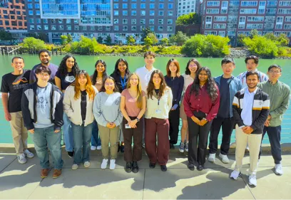 MSBI students standing together outdoors along a waterfront, posing for a group photo with campus buildings in the background.
