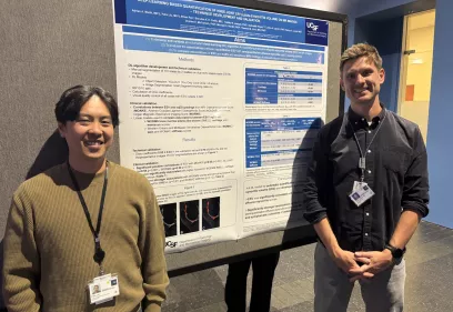 Two researchers stand on either side of a large scientific poster titled “Deep-Learning Based Quantification of Knee Joint Effusion–Synovitis Volume on MR Images: Technique Development and Validation” displayed on a poster board at a conference. Both are wearing lanyards with identification badges. The researcher on the left wears a tan sweater and smiles toward the camera, while the researcher on the right wears a dark button-up shirt and stands with hands clasped. The poster includes sections such as Aims
