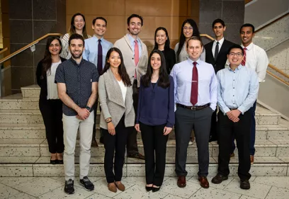 Portrait of a group of first-year medical residents and a faculty member standing on a staircase indoors. The group is dressed in business attire and posed in three rows, smiling toward the camera.
