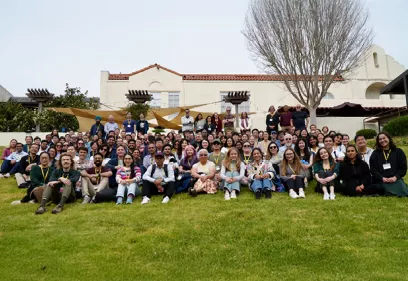 Large group photo of attendees seated and standing on a grassy lawn in front of a building during a research conference.