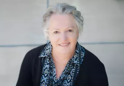 Portrait of a smiling older woman with short gray hair, wearing a patterned blouse and dark cardigan, against a soft neutral background.