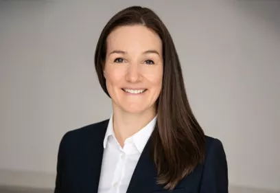 Professional headshot of a woman smiling at the camera. She has long, straight dark brown hair parted to one side and is wearing a dark navy blazer over a white collared shirt. The portrait is framed from the chest up against a soft, light gray background. The lighting is even and natural, creating a polished and approachable appearance suitable for a professional, academic, or corporate profile.