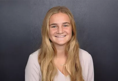 Portrait of Allison smiling at the camera against a dark gray background. She has long blonde hair and is wearing a light-colored blouse and a simple necklace.