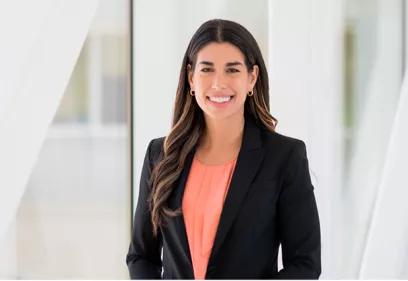 Professional portrait of a woman with long dark wavy hair wearing a black blazer over a coral blouse. She is smiling at the camera while standing in a bright, modern indoor space with large windows and softly blurred white architectural elements in the background.