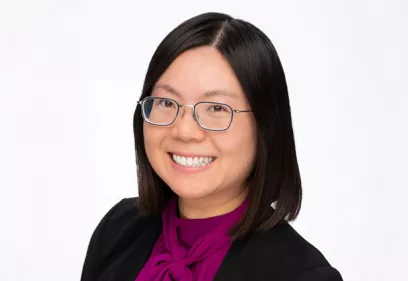 Professional headshot of a smiling woman with shoulder-length dark hair and glasses, facing the camera. She is wearing a black blazer over a magenta blouse with a tied neckline. The portrait is set against a light gray background, creating a clean and polished appearance suitable for a professional or faculty profile.